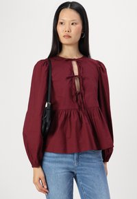 Bordo peplum bluzonė su pūstomis rankovėmis ir skylutės detale iškirptėje. Medvilninė medžiaga su lygiu paviršiumi, derinama su mėlynais džinsais.