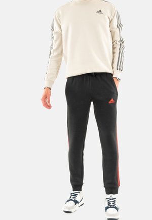 Sudadera beige de Adidas con rayas grises en las mangas, combinada con pantalones deportivos negros que tienen acentos rojos en los laterales. Zapatillas deportivas blancas y marinas.