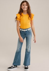 Dzeltena ribota T-kreklu ar volāniem uz piedurknēm, apvienota ar ziliem platām biksēm un melniem sporta apaviem ar baltām akcentiem.