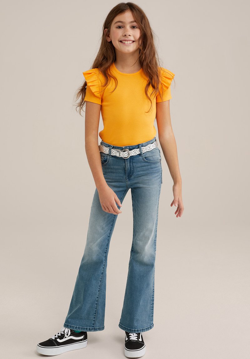 Dzeltena ribota T-kreklu ar volāniem uz piedurknēm, apvienota ar ziliem platām biksēm un melniem sporta apaviem ar baltām akcentiem.