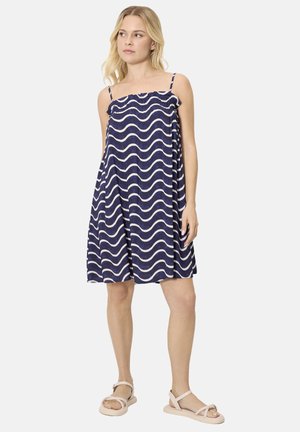 Mujer rubia con vestido azul marino sin mangas con patrón de olas blancas y sandalias beige, de pie con los brazos relajados, mirando hacia la izquierda.