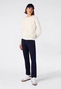 Felpa color crema con scollo rotondo, abbinata a pantaloni blu navy a sigaretta e sneakers bianche. Il modello indossa un berretto nero.