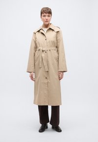 Ταν παλτό καμπαρντίνα με ζώνη στη μέση, μεγάλο γιακά και κουμπιά κατά μήκος του μπροστινού μέρους. Διαθέτει μακριά μανίκια και ευθύγραμμη σιλουέτα.