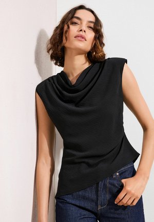 Žena s krátkymi kučeravými vlasmi v čiernom asymetrickom tielku bez rukávov a modrých džínsach, opierajúca sa o bielu stenu.