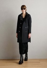 Manteau noir croisé avec de grands boutons, large col, et poches latérales ; porté sur une jupe noire texturée et des bottes à plateforme noires épaisses.