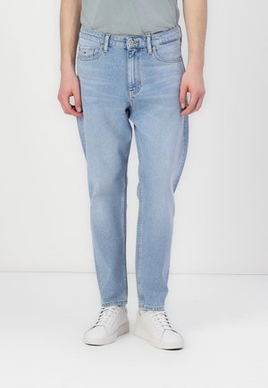 Mann trägt hellblaue Jeans und weiße Turnschuhe, steht vor einem schlichten weißen Hintergrund.