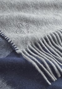 Grau Wolldecke mit Fransen, die ein hellgraues besticktes Blumendesign zeigt, übereinandergelegt auf einem dunkelblauen Hintergrund.