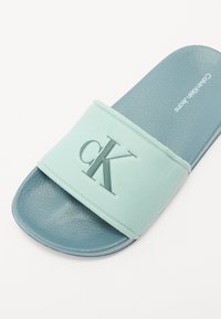 Himmelblauer Rutschsandale mit einer strukturierten Sohle. Verfügt über einen weichen, mintgrünen Riemen, der mit "cK" in passender Farbe geprägt ist.