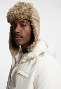 Beige nep bont gevoerde muts met oorkleppen, gemaakt van zachte stof. Draagt een witte jas met rits en schapenvachtkraag.