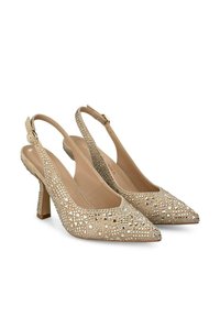 Zapatos de tacón alto beige tipo slingback con puntera puntiaguda, decorados con tachuelas plateadas geométricas y un tacón texturizado. Incluye correa ajustable para el tobillo.