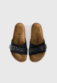 Sandales en suede noir avec une sangle tressée et une boucle rectangulaire en argent. Semelle en liège offrant une finition naturelle. Design à semelle plate.