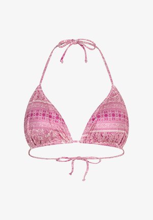 Frau mit langem Haar, die einen rosa gemusterten Bikini mit Bindeseiten trägt, steht drinnen neben einer Holztür und einer grünen Pflanze.