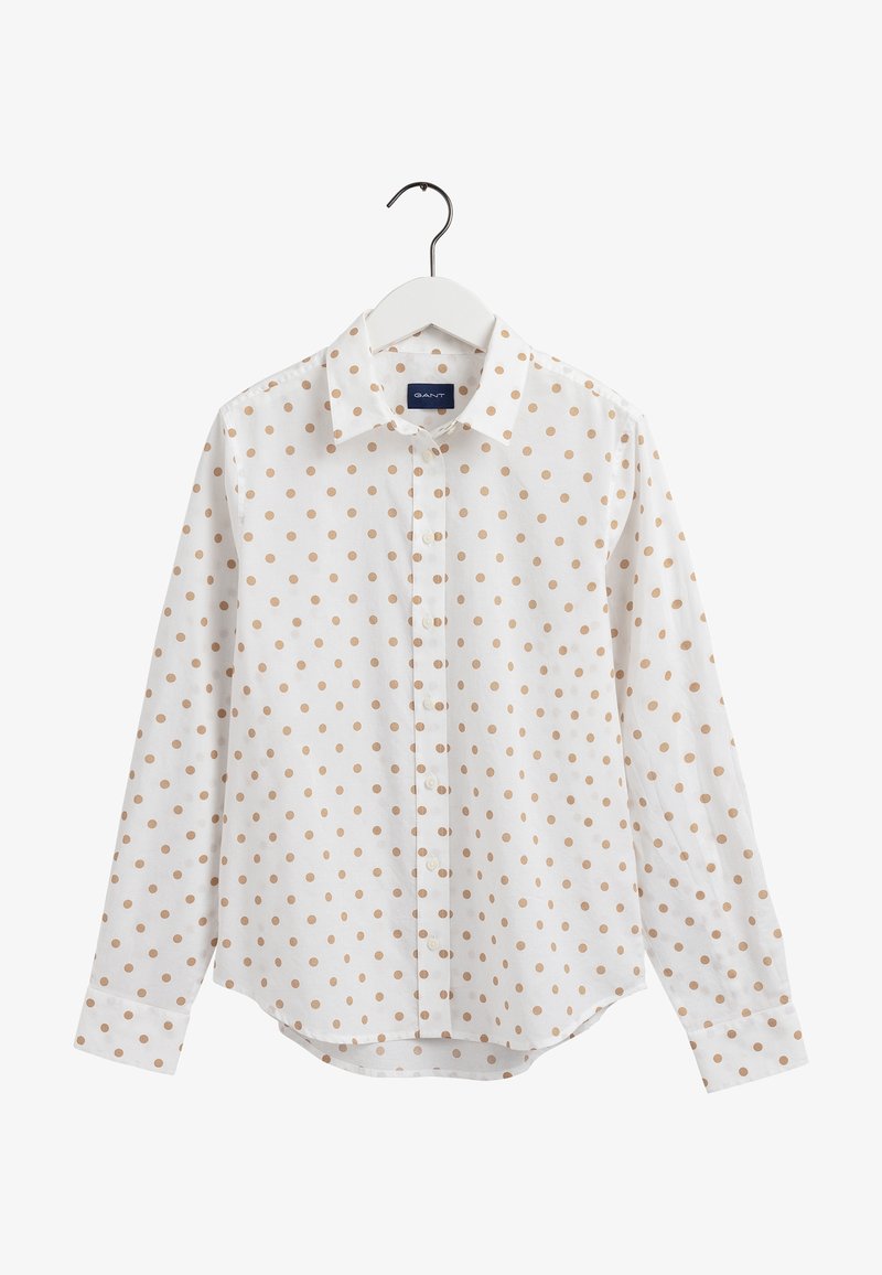 Chemise blanche à manches longues avec des pois beiges, présentée sur un cintre blanc sur un fond uni.