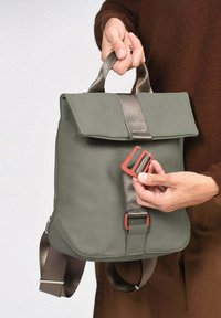 Sac vert imperméable avec rabat, doté d'une boucle avant et d'un tissu résistant. Des bretelles réglables et une poche latérale sont visibles.