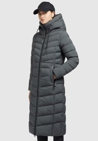 Manteau rembourré gris avec un design long et matelassé, un col montant et une capuche ajustable. Il dispose de poches latérales et d'une fermeture éclair à l'avant. Les accents incluent des étiquettes de logo.