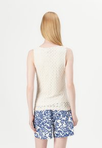 Strikket ermeløs topp i krem med teksturert mønster; kombinert med blå og hvite blomstrete shorts. Minimalistisk design og lett materiale.