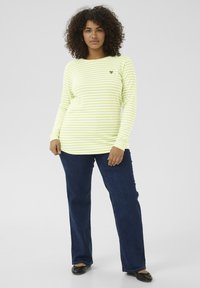 T-shirt à manches longues, rayé jaune clair et blanc avec un petit emblème en forme de cœur sur la poitrine, associé à un jean bleu foncé et des ballerines noires.