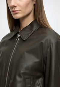 Casaco de couro castanho escuro com gola pontiaguda e fecho prateado. Apresenta uma textura lisa e ombros estruturados, exibido em perfil lateral.