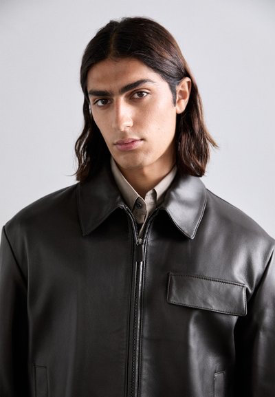 Brown leather jacket featuring a collared neck, zip closure, and a patch pocket on the left side. Smooth texture with minimal stitching details.