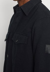 Uomo che indossa un giacca in lana blu scuro con bottoni, dotata di tasche sul petto e colletto, mostrato dalla nuca fino alla parte centrale del torace.