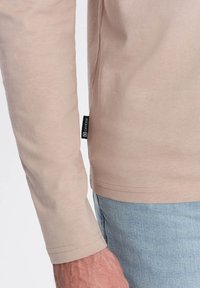 Långärmad beige skjorta i mjukt tyg, med en etikett på sidan. Ljusblå denimjeans syns i kombination.