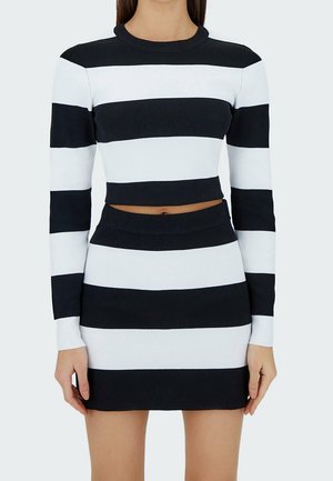 Femme portant un crop top à manches longues rayé horizontalement noir et blanc et une mini jupe assortie sur un fond uni.