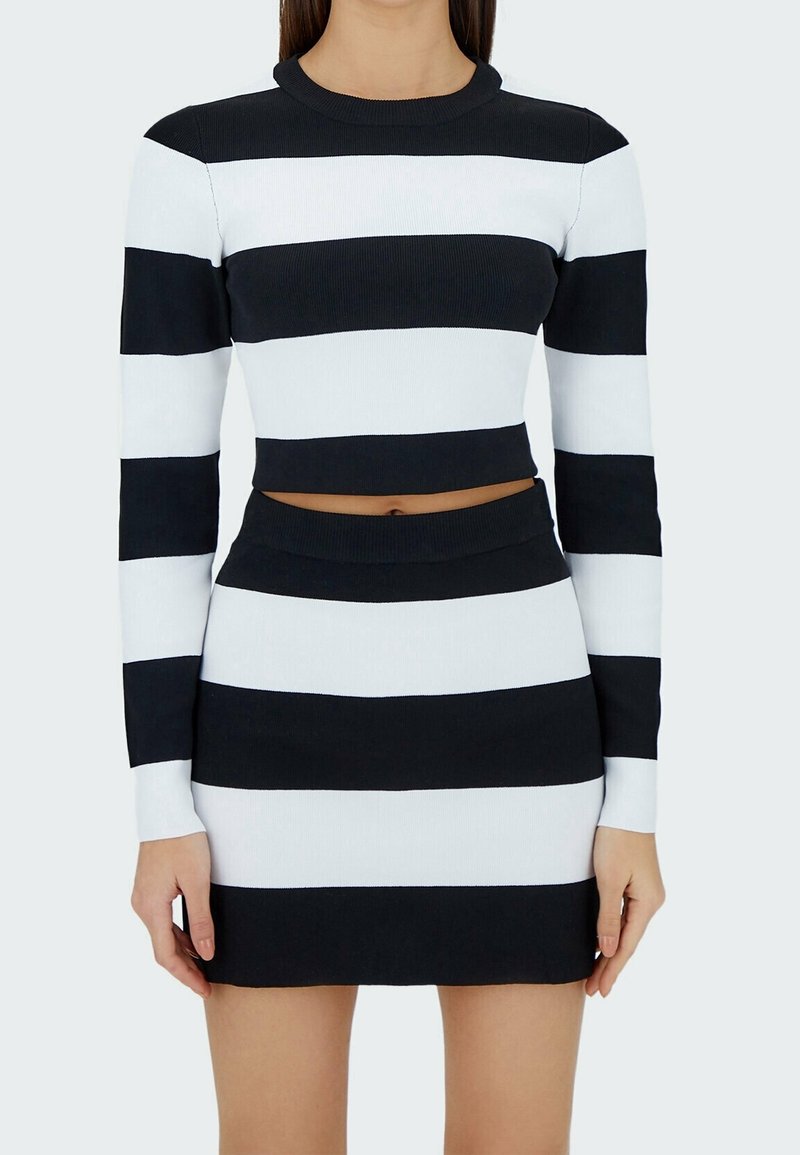 Femme portant un crop top à manches longues rayé horizontalement noir et blanc et une mini jupe assortie sur un fond uni.
