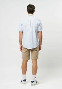 Lichtblauwe blouse met korte mouwen en een plooitje aan de achterkant, gecombineerd met beige shorts en witte sokken, gedragen met casual sneakers.