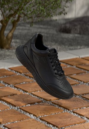 Sneaker in pelle nera con lacci, sospesa sopra una piastrella di legno e una superficie esterna di ghiaia con un piccolo albero sullo sfondo.