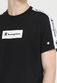 Černé bavlněné tričko s bílým logem "Champion" ve středu a bílou značkovou páskou podél ramenních švů. Krátké rukávy.