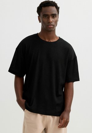 Joven con cabello corto y rizado que lleva una camisa negra suelta de manga corta y pantalones beige sobre un fondo blanco liso.