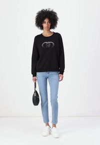 Sudadera negra con cuello redondo y diseño impreso, combinada con jeans de un azul claro y zapatillas blancas. Un pequeño bolso negro completa el look.