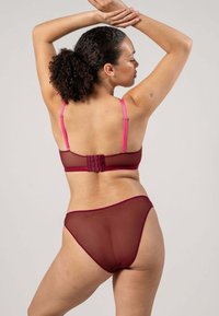 Kvinna med lockigt hår i hästsvans iförd en genomskinlig bordeauxfärgad BH och trosor, poserar med armarna uppsträckta mot en enkel bakgrund.