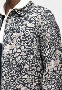 Mężczyzna ubrany w czarno-białą pikowaną kurtkę z abstrakcyjnym kwiatowym wzorem, na białej koszuli z kołnierzykiem i widocznym zamkiem błyskawicznym.