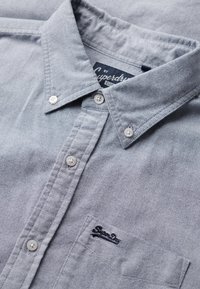 Lichtgrijze button-up shirt gemaakt van stof met een subtiele textuur. Bevat een kraag, twee knopen bij de hals en een borstzak met branding.