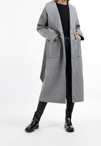 Long manteau gris en laine avec grandes poches, large col et boutons à l'avant, porté sur un haut noir et un jean foncé avec des bottines noires brillantes.