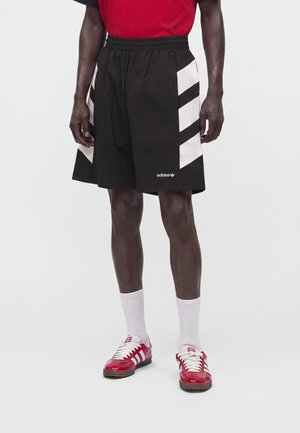 Osoba nosiaca čierne šortky Adidas s bielymi pruhmi po stranách, biele ponožky a červené tenisky Adidas s bielymi šnúrkami.