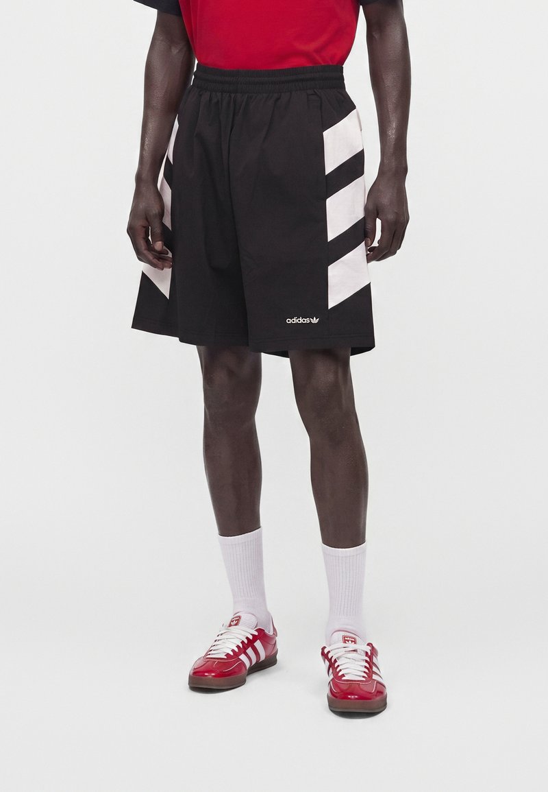 Άτομο που φοράει μαύρο σορτς Adidas με λευκές ρίγες στα πλευρά, λευκές κάλτσες και κόκκινα αθλητικά παπούτσια Adidas με λευκά κορδόνια.