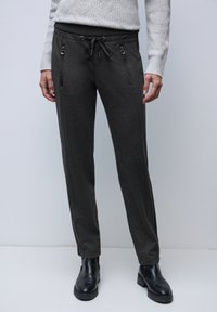 Person trägt dunkelgraue Kordelzughose mit Reißverschlusstaschen und schwarze Stiefeletten, kombiniert mit einem hellgrauen, strukturierten Pullover.