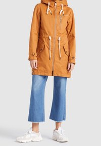 Pomarańczowa wodoodporna kurtka parka z kapturem na sznurek, zamkiem błyskawicznym i przednimi kieszeniami. W komplecie z jasnoniebieskimi szerokimi spodniami i białymi tenisówkami.