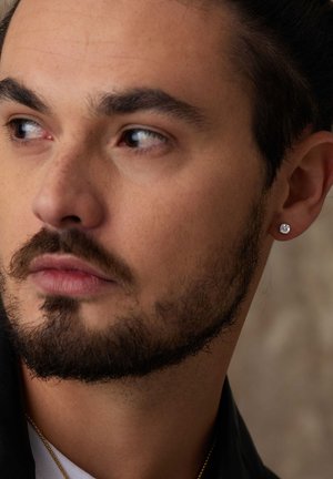 Boucle d'oreille en argent avec une pierre précieuse transparente, portée à l'oreille gauche. Gros plan sur la boucle d'oreille et les traits du visage, fond neutre.