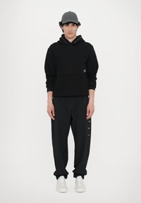 Felpa nera con tasca frontale e logo, abbinata a pantaloni neri larghi con un logo stampato sul lato e sneakers bianche.