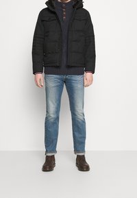 Mann, der eine schwarze gepolsterte Jacke, einen navyblauen Strickpullover, hellblaue hochgerollte Jeans und braune Leder-Stiefel bis zum Knöchel trägt, steht vor einem weißen Hintergrund.