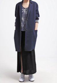 Námořnický dlouhý blazer se dvěma velkými předními kapsami, nošený přes vzorovaný top a černou sukni, doplněný bílými teniskami pro ležérní vzhled.