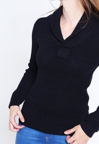 Navy pletený svetr s límcem typu šál, dlouhými rukávy a texturovaným materiálem. Obsahuje malý knoflíkový detail na límci a je přiléhavého střihu.