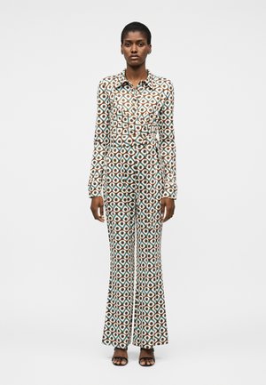 Γυναίκα που φορά ολόσωμη φόρμα με μακριά μανίκια και γεωμετρικό σχέδιο, με παντελόνια σε ευρύ στυλ και μαύρα σανδάλια με ανοιχτά δάχτυλα, στέκεται μπροστά σε απλό φόντο.