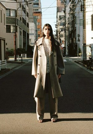 Vrouw staand in een stadsstraat, gekleed in een lange beige jas, een wit gestructureerd topje en wijde broekspijpen, met gebouwen en elektriciteitskabels op de achtergrond.