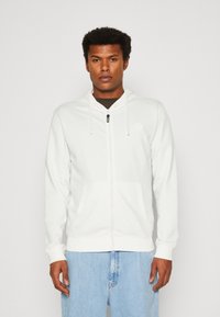 Bijela zip-up dukserica od mekog materijala, s diskretnim logom, kapuljačom na vezivanje i dva prednja dzepa. Uparena s svijetloplavim farmerkama.