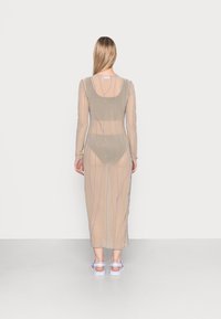 Vestido longo de mangas largas em tom bege transparente, com costura irregular em roxo, costas em escopo e silhueta ajustada, combinado com sandálias de tom claro.