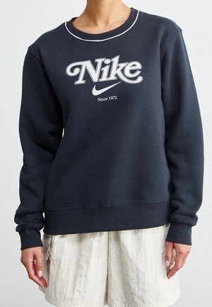 Personne portant un sweat-shirt Nike bleu marine foncé avec un logo blanc et un texte "Since 1972,", associé à un short texturé blanc cassé.